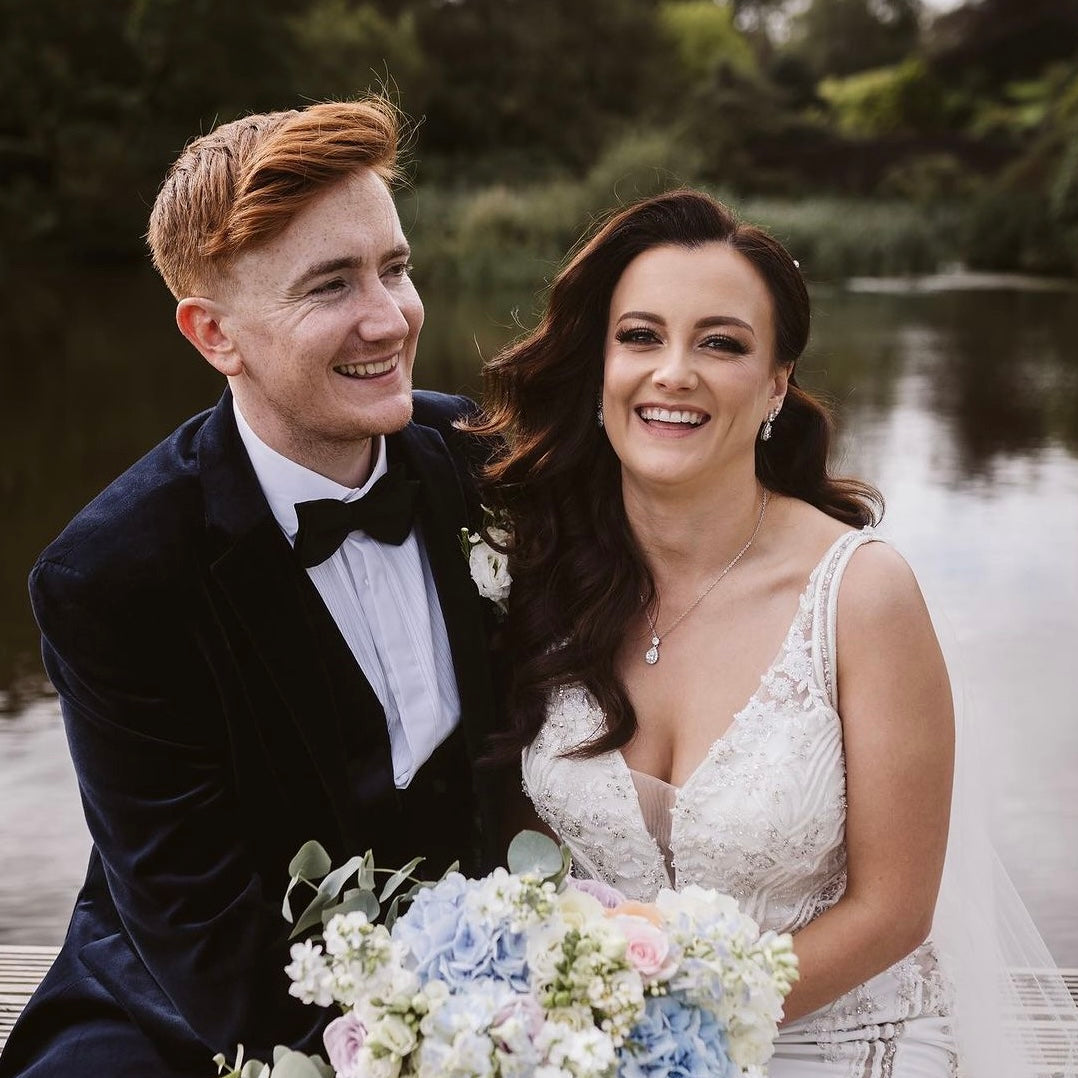 A smiling bride and groom sitting together on a wooden dock by a lake. The groom wears a dark velvet tuxedo jacket with a white shirt and black bow tie, and the bride wears a white lace wedding gown and holds a bouquet of white, blue, and peach flowers.