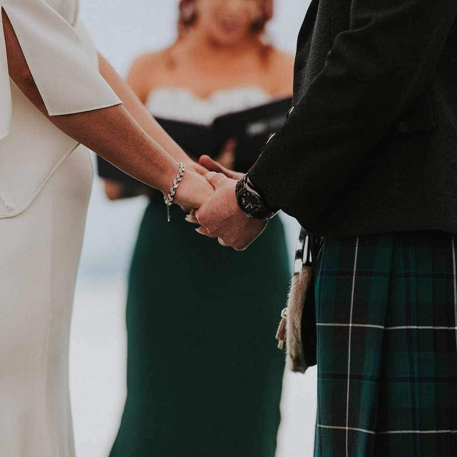 A close-up wedding scene showing two people holding hands outdoors. One person wears an off-white sleeveless dress, and the other wears a dark blazer and a green tartan kilt.